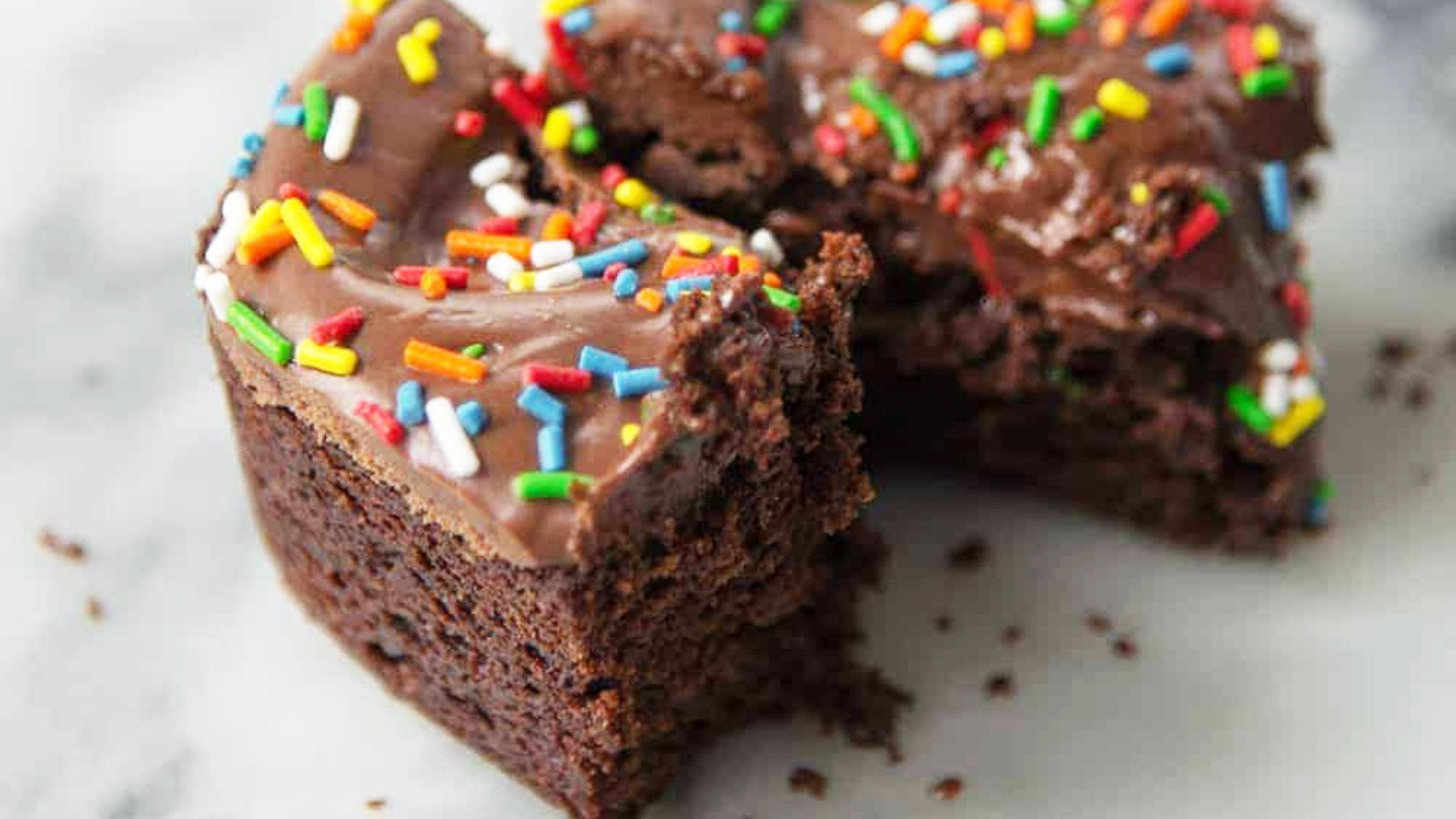 A closeup of a cut brownies with frosting and colored sprinkles on a white surface.