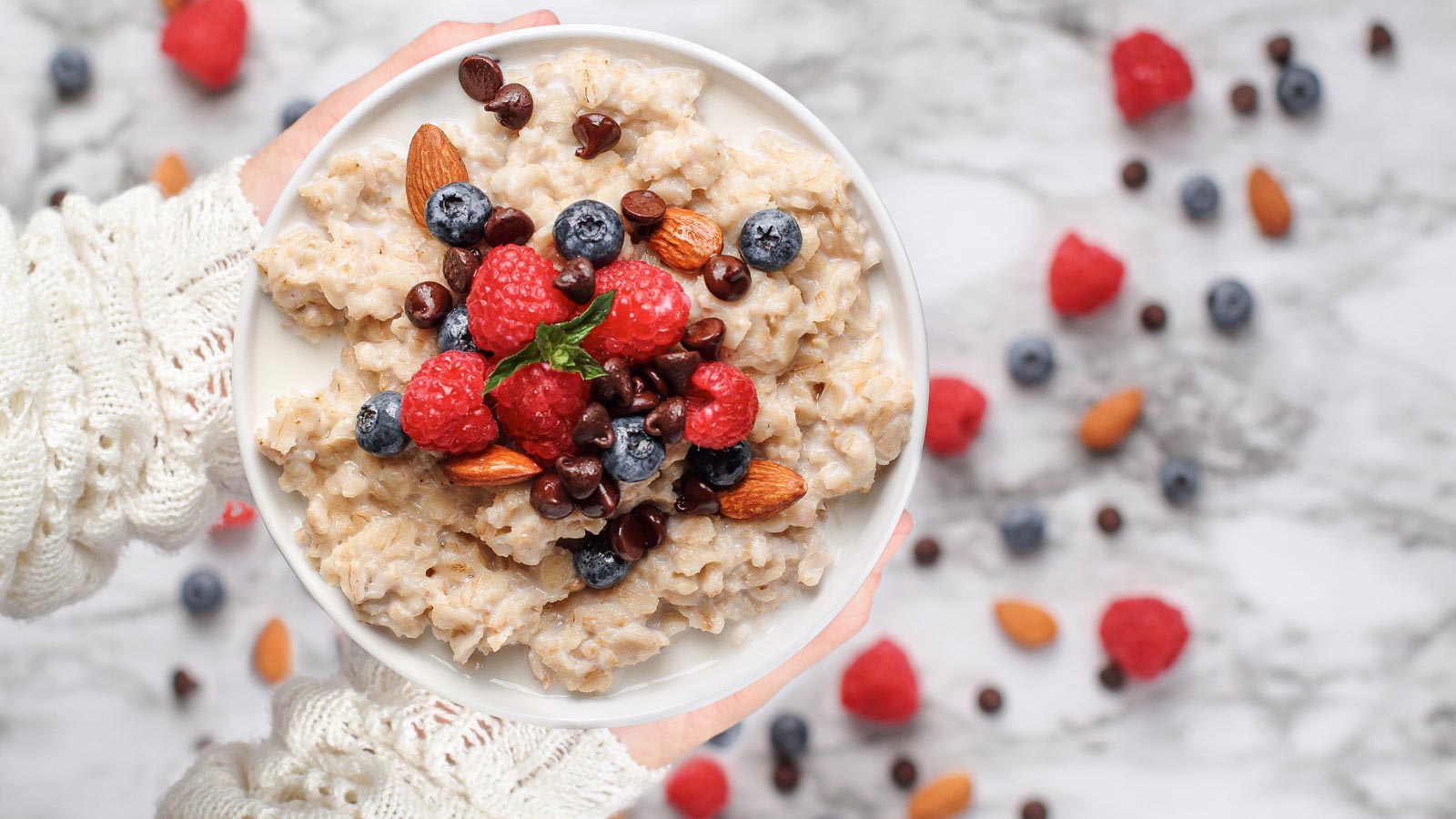 Almond meal porridge served with berries, chocolate chips, almonds and honey. Bowl held in a womans hands over a marble table background. Shot from top view.