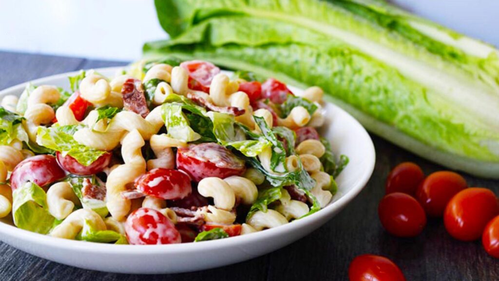 BLT Pasta Salad in a white bowl with a head of lettuce and some grape tomatoes sitting behind the bowl.
