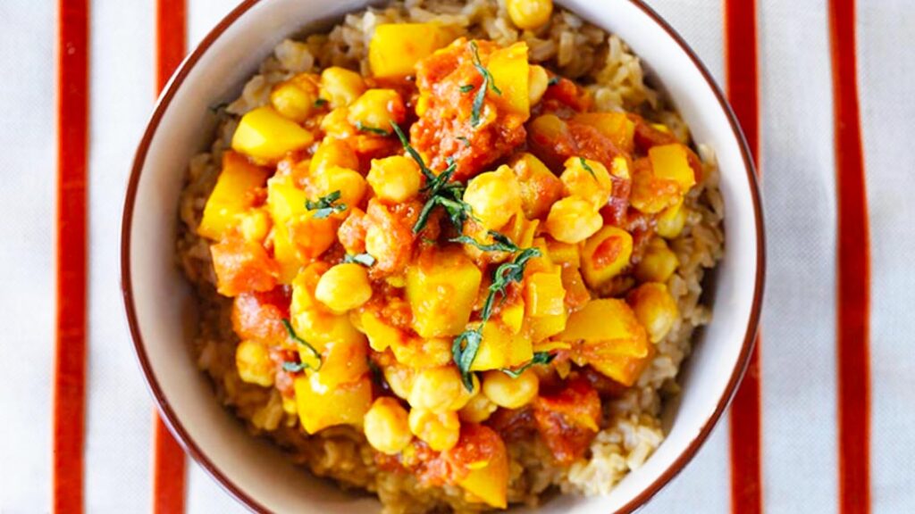 An overhead view of a bowl of Bombay potatoes in a bowl on a striped tablecloth.