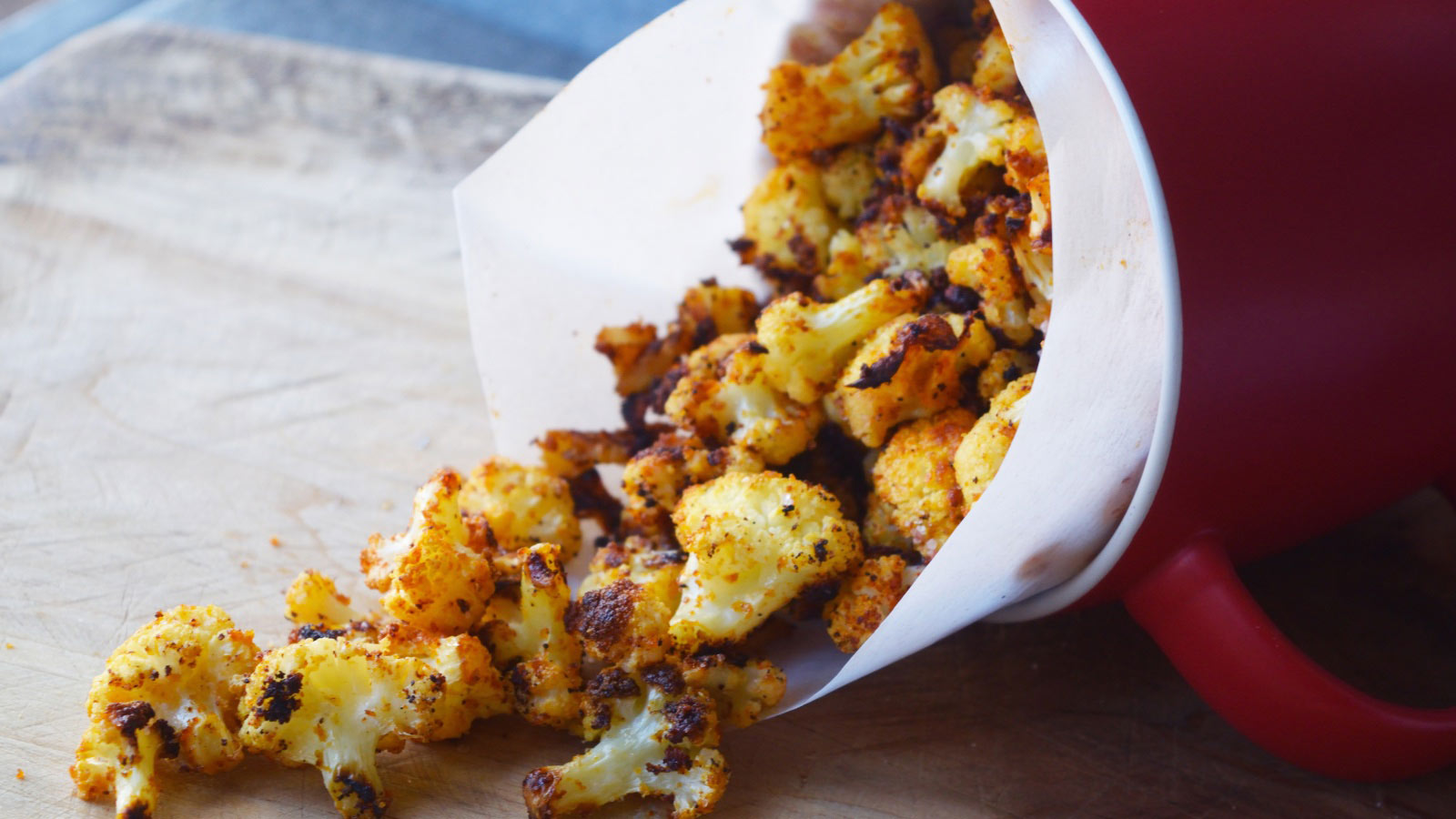 A red mug, tipped over, with Cauliflower Popcorn cascading out of it onto a wood cutting board.