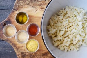 A bowl of small cauliflower pieces next to several small bowls of spices and oil.