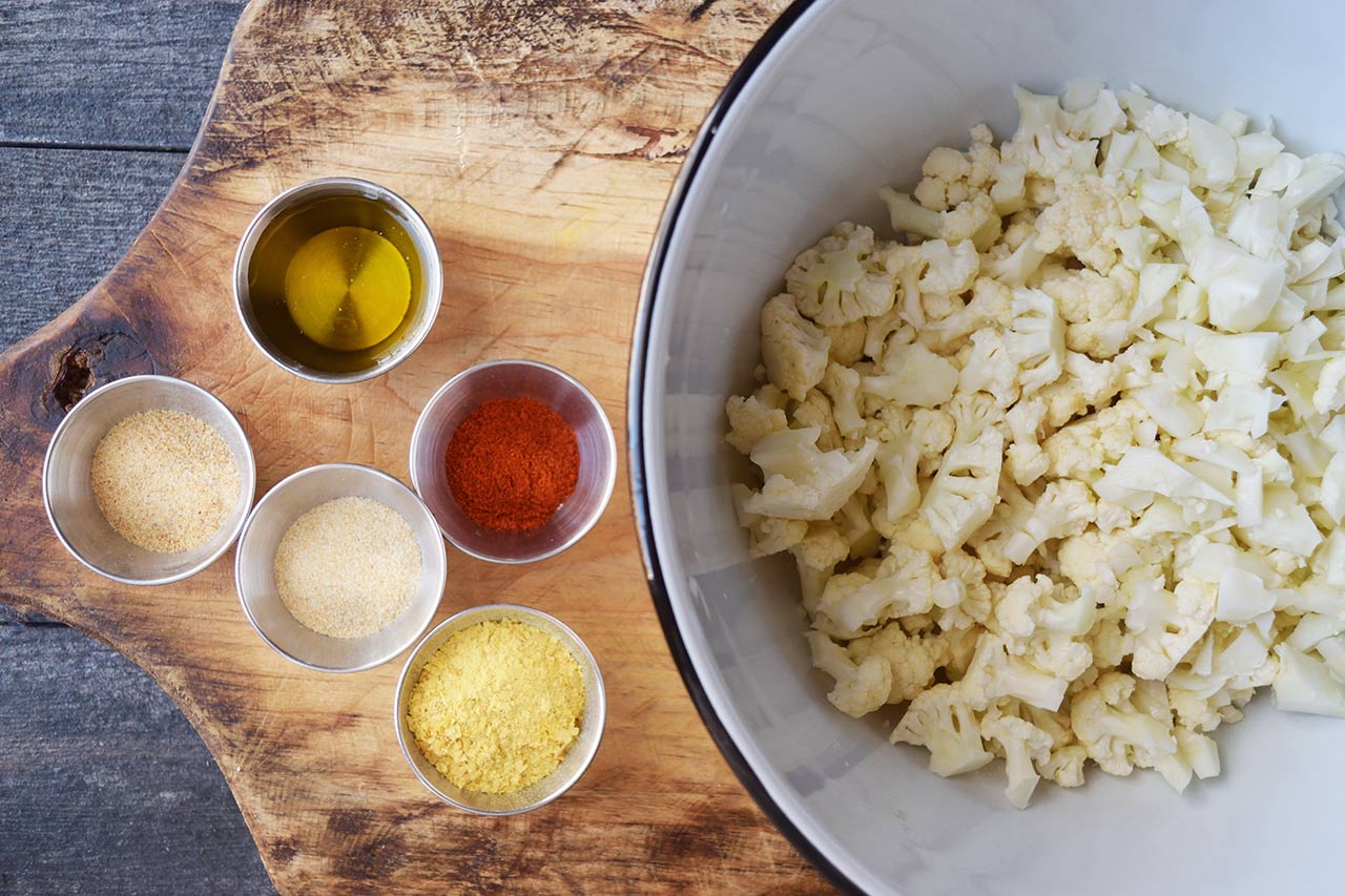 A bowl of small cauliflower pieces next to several small bowls of spices and oil.