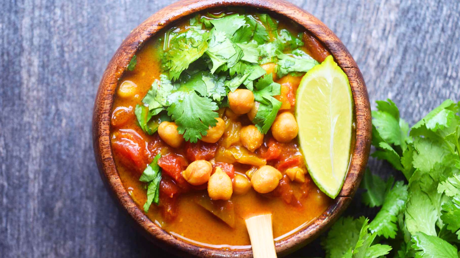 An overhead view of a wood bowl filled with Chickpea Curry and garnished with fresh, chopped cilantro and a lime wedge.