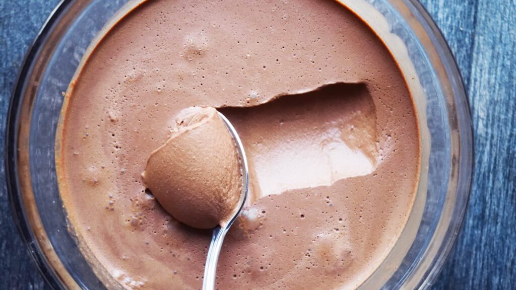 A spoon scoops up some chocolate mousse from a glass bowl full of mousse.
