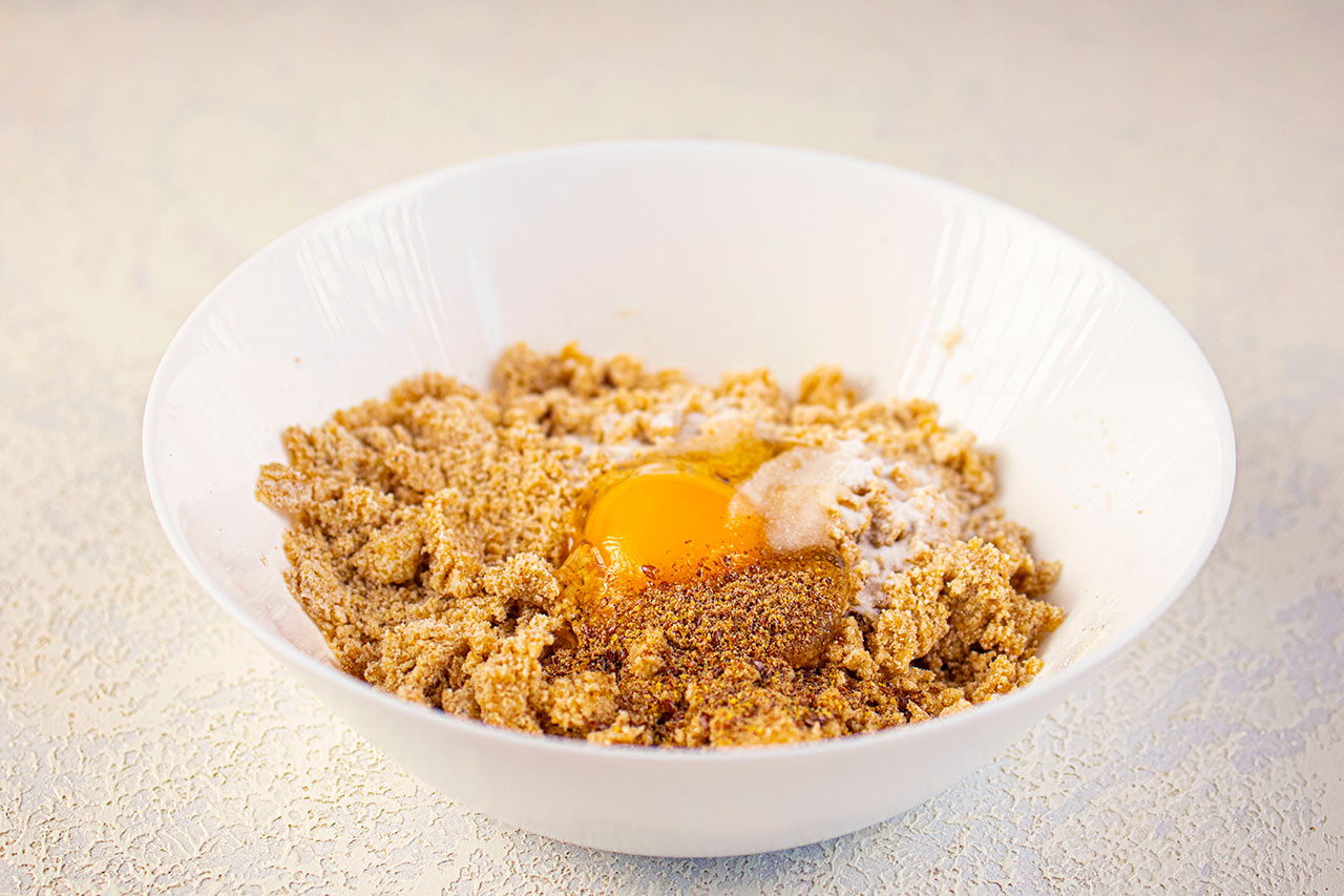 An egg added to the graham cracker mix in a white mixing bowl.