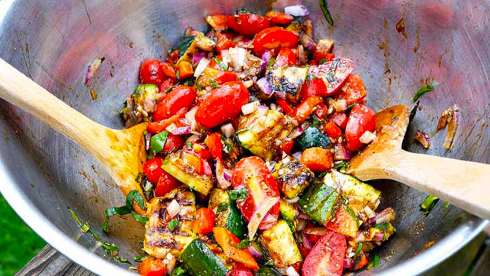 A side view of a metal bowl with wooden tongs, partially filled with grilled zucchini salad.