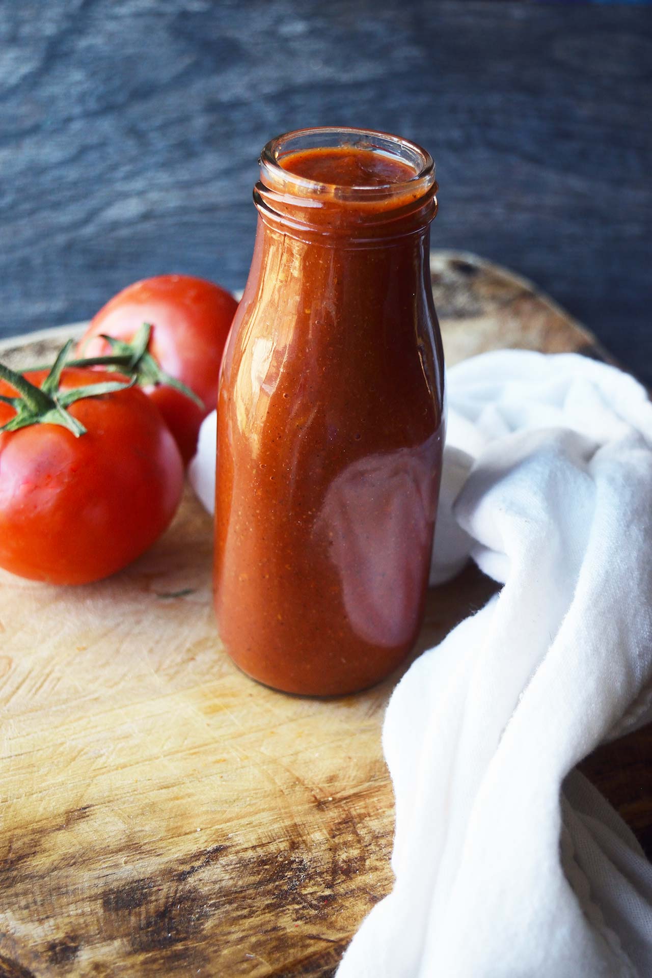 Ketchup in a glass jar sitting on a wood cutting board with two tomatoes and white towel.