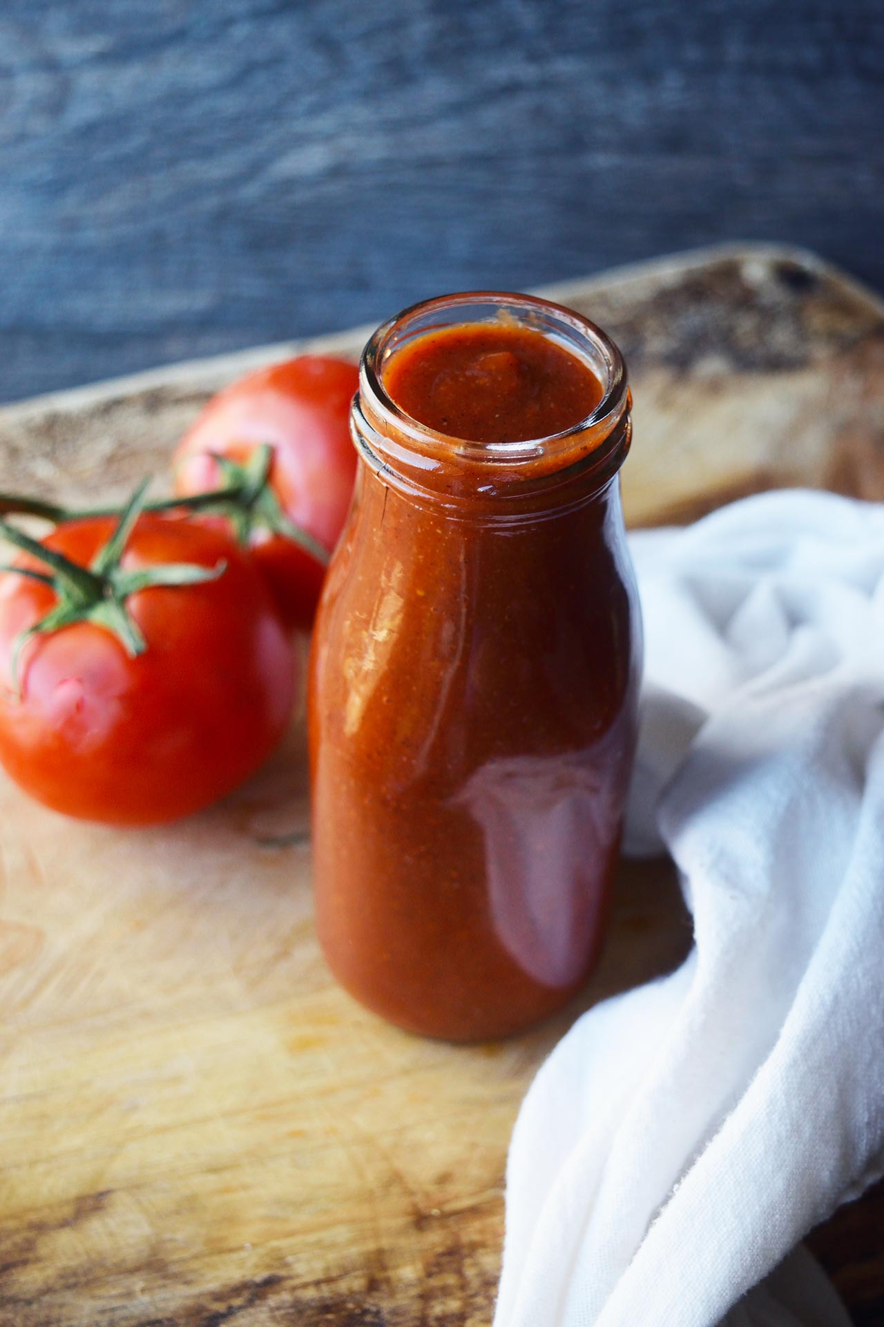 A side view of a glass bottle of homemade ketchup sitting on a wooden cutting board.