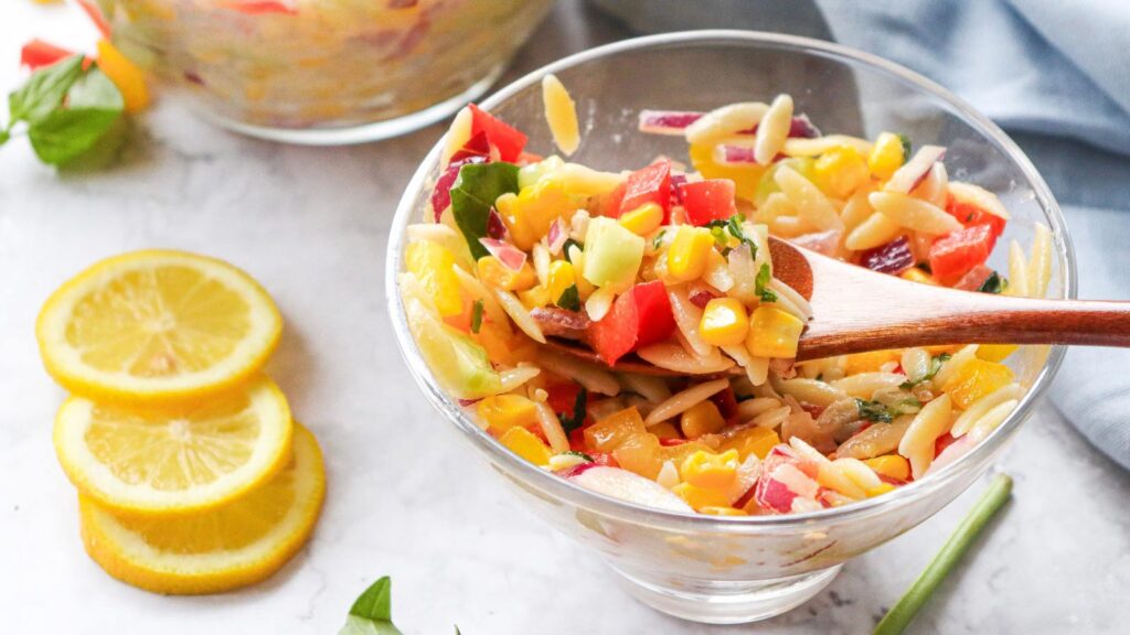 A glass bowl filled with Rainbow Orzo Salad with a wooden spoon resting in it.