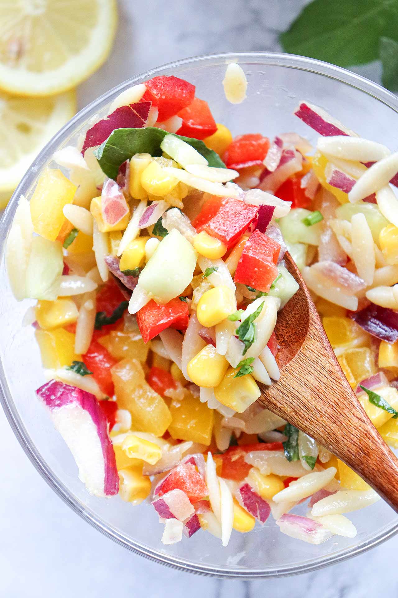 A wooden spoon lifting a spoonful of rainbow orzo salad from a glass bowl.