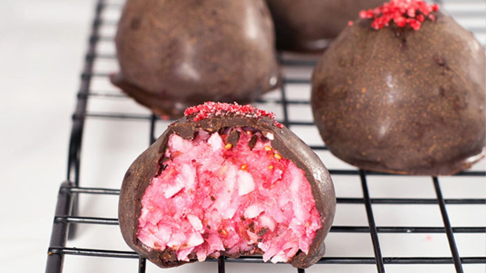 Just cooled Raspberry Coconut Truffles sitting on a cooling rack. The front truffle has a bite taken from it.