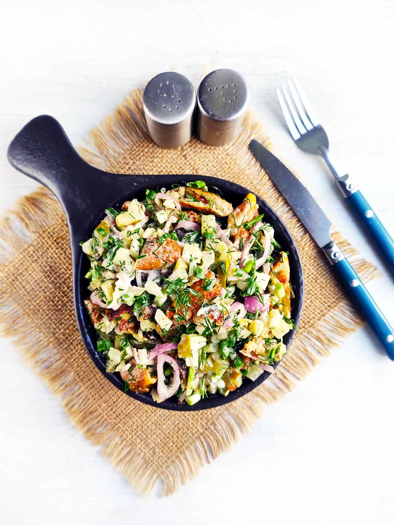 An overhead view of a black bowl filled with Red Potato Salad, sitting on a placemat with a knife, fork, salt, and pepper shaker.