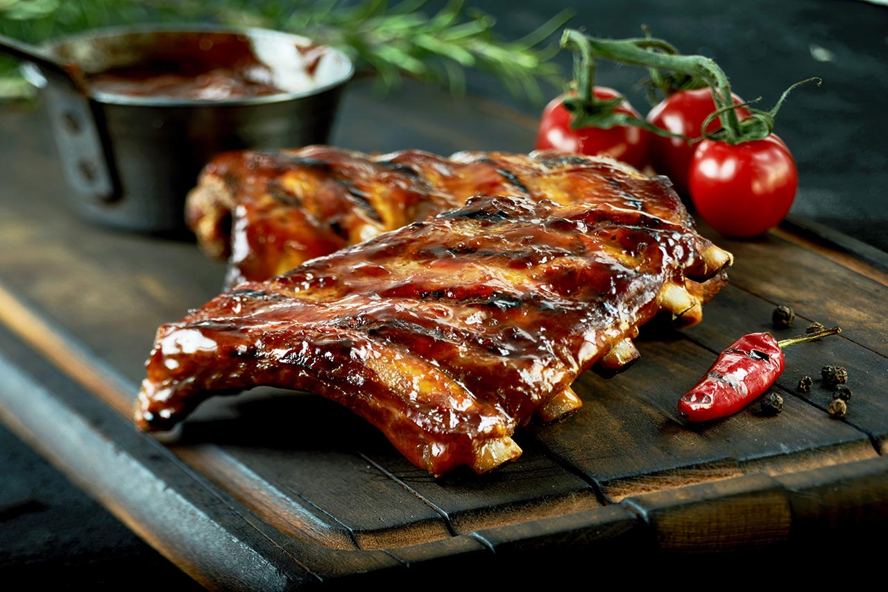 Spicy hot grilled spare ribs from a summer BBQ served with a hot chili pepper and fresh tomatoes on an old vintage wooden cutting board.