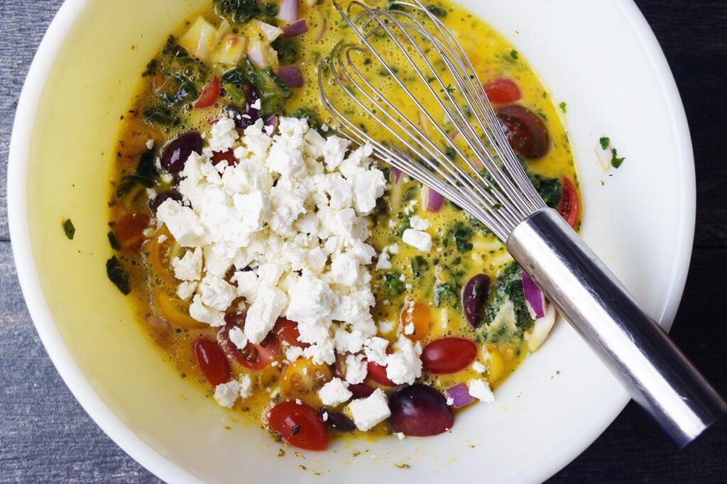 Quiche ingredients and additions added to the whisked eggs and spices in a mixing bowl.