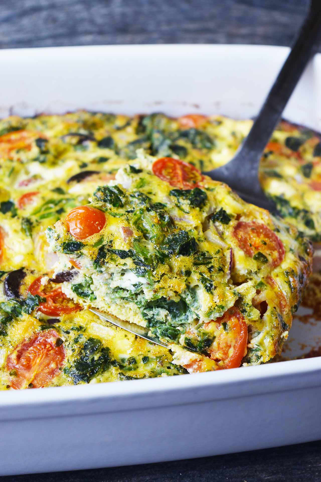 A metal spatula holds a serving of spinach quiche over the casserole dish it was taken from.