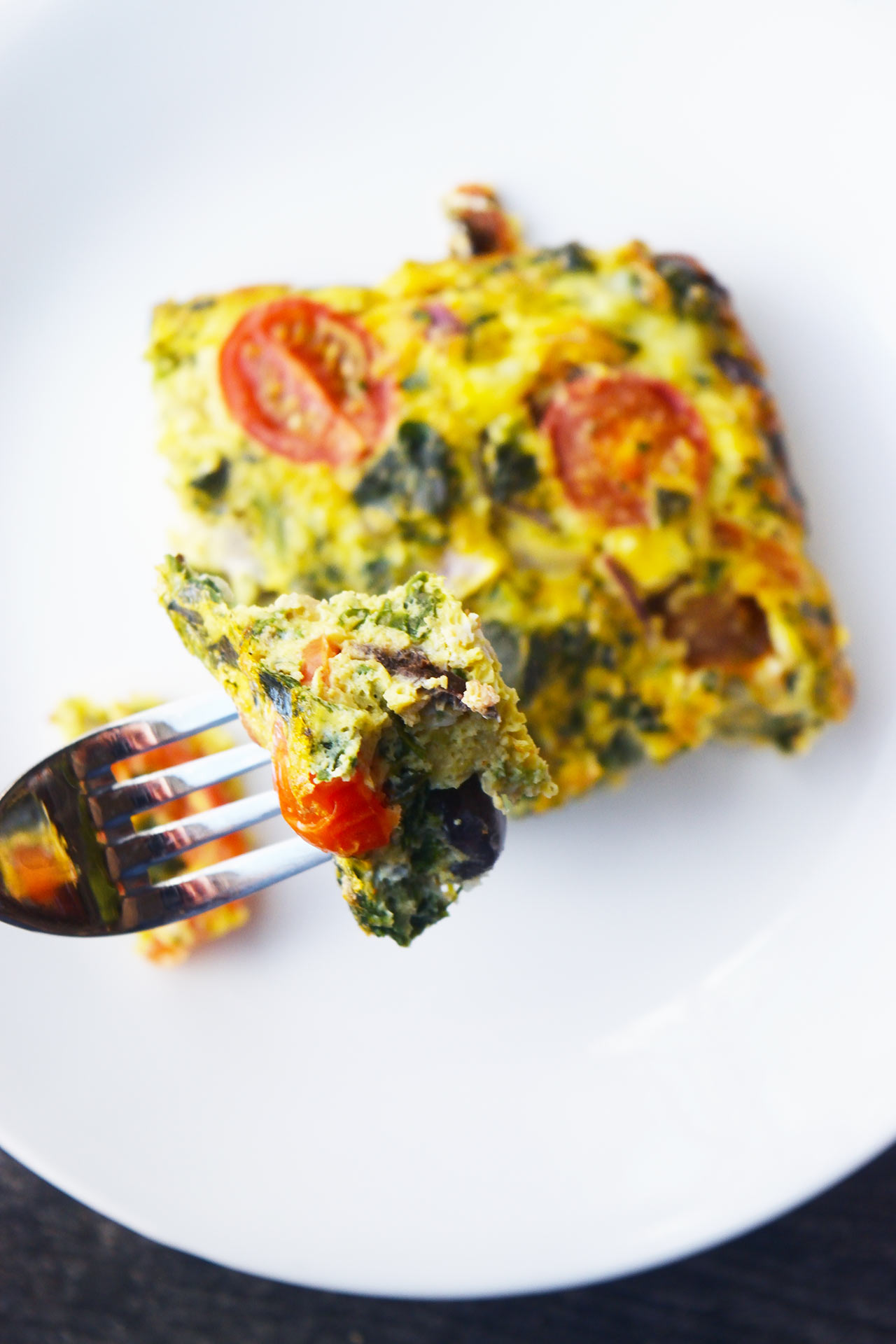 An overhead view of a piece of spinach quiche on a white plate. A fork lifts a bite of the quiche from the plate.