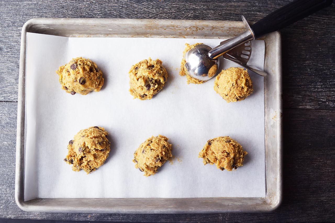 A cookie scoop rests on a baking sheet with six scoops of cookie dough lined up for baking.