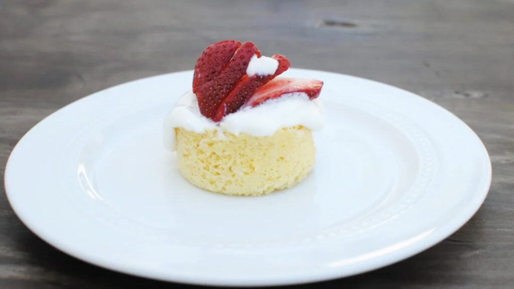 A single mug cake on a plate, garnished with strawberry slices and whipped cream.