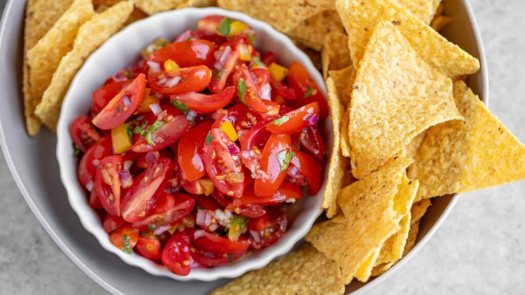 An overhead view of a white bowl filled with cherry tomatoes salsa and surrounded by corn chips.