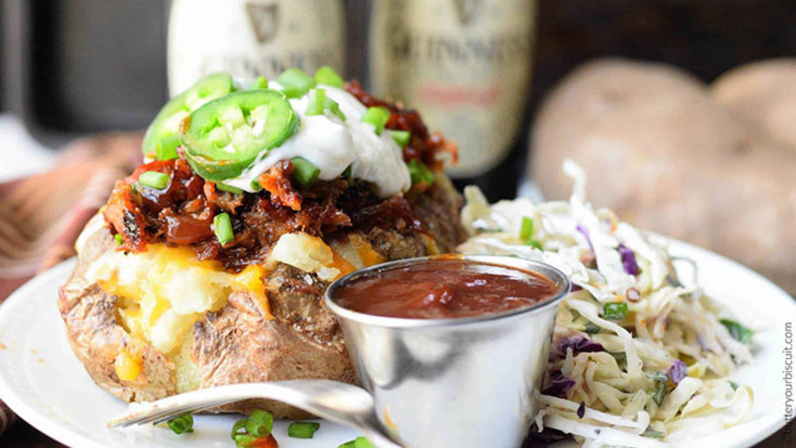 A pulled pork loaded baked potato on a white plate with coleslaw and a small metal cup of sauce.