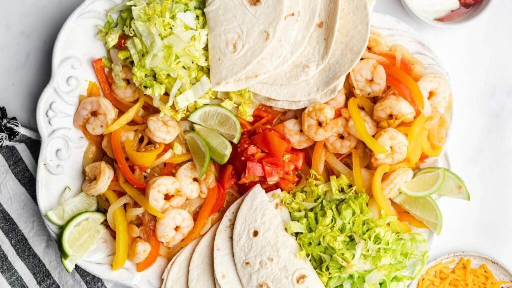 A platter with shrimp, veggies, lettuce, and tortillas for assembling shrimp fajitas.