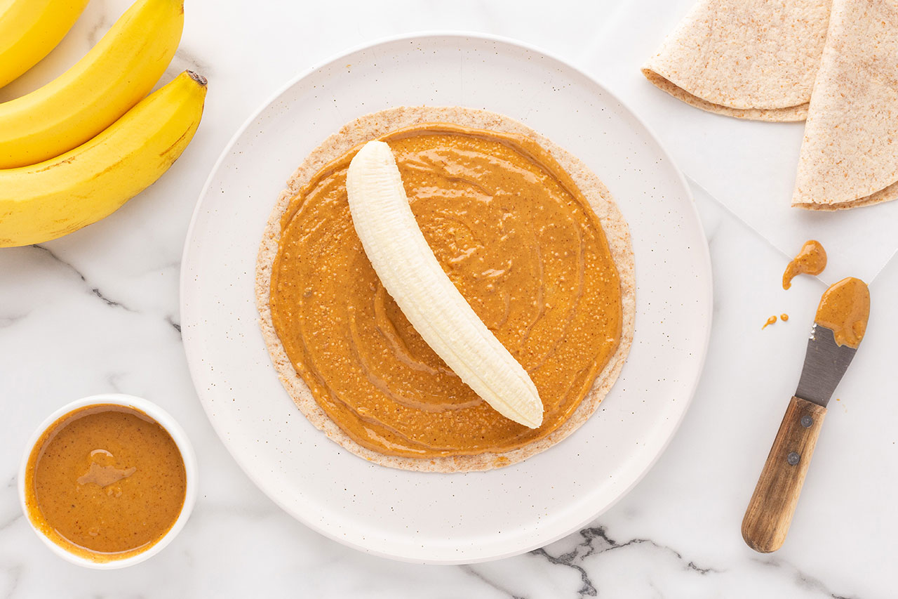 A banana placed in the center of a tortilla covered in peanut butter laying on a white plate.