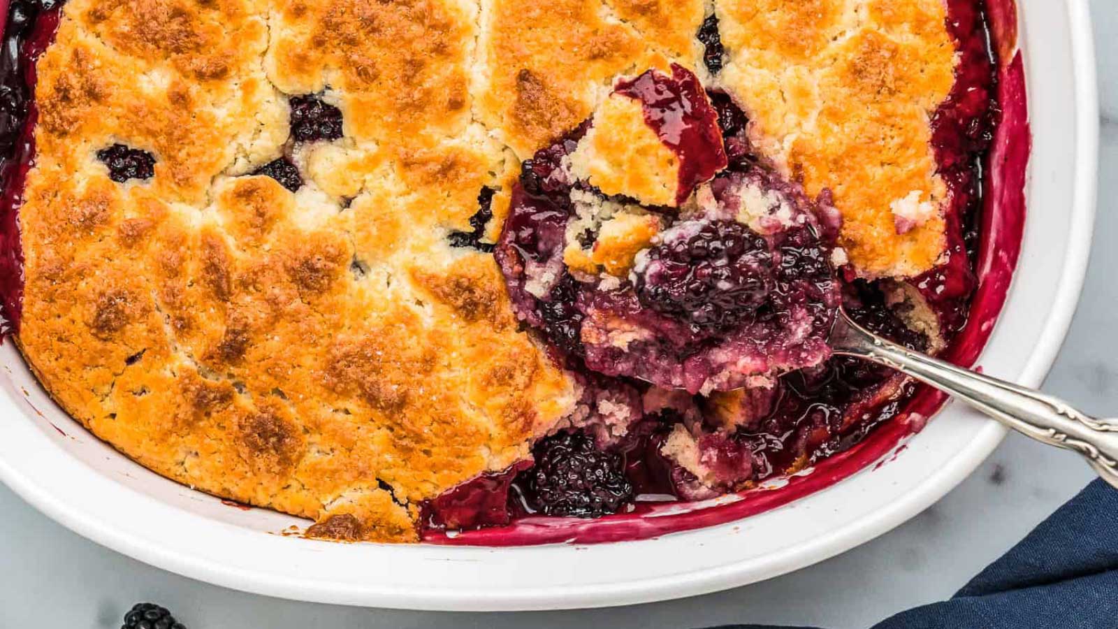 An overhead view of a white casserole dish filled with black berry cobbler and a serving spoon.