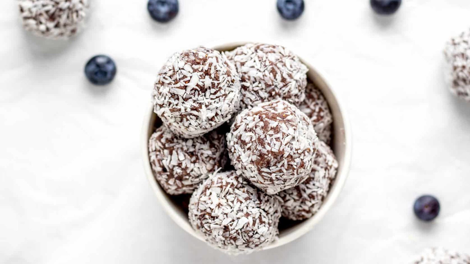 An overhead view of a white bowl on a white surface. The bowl is filled with blueberry bliss balls and there are fresh blueberries sprinkled around the bowl.