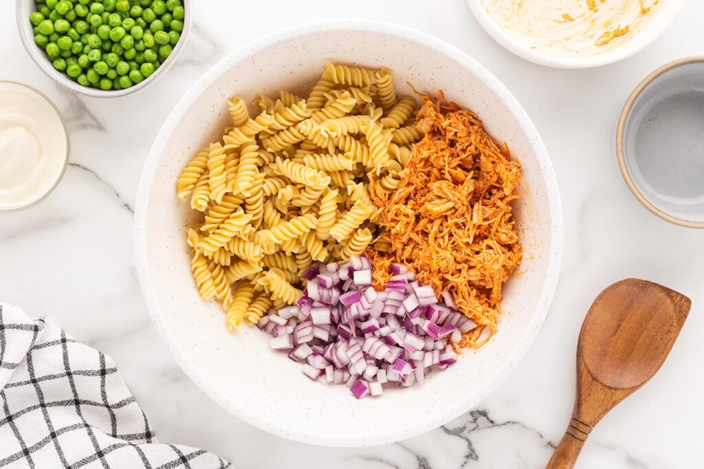 Chopped onions added to a mixing bowl with buffalo chicken and pasta.