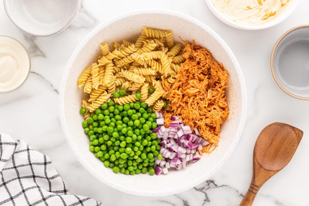 Pease added to a mixing bowl with pasta, buffalo chicken, and onions.