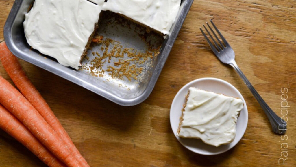 A pan of cut carrot cake with one slice on a small plate to the right. Overhead focus.