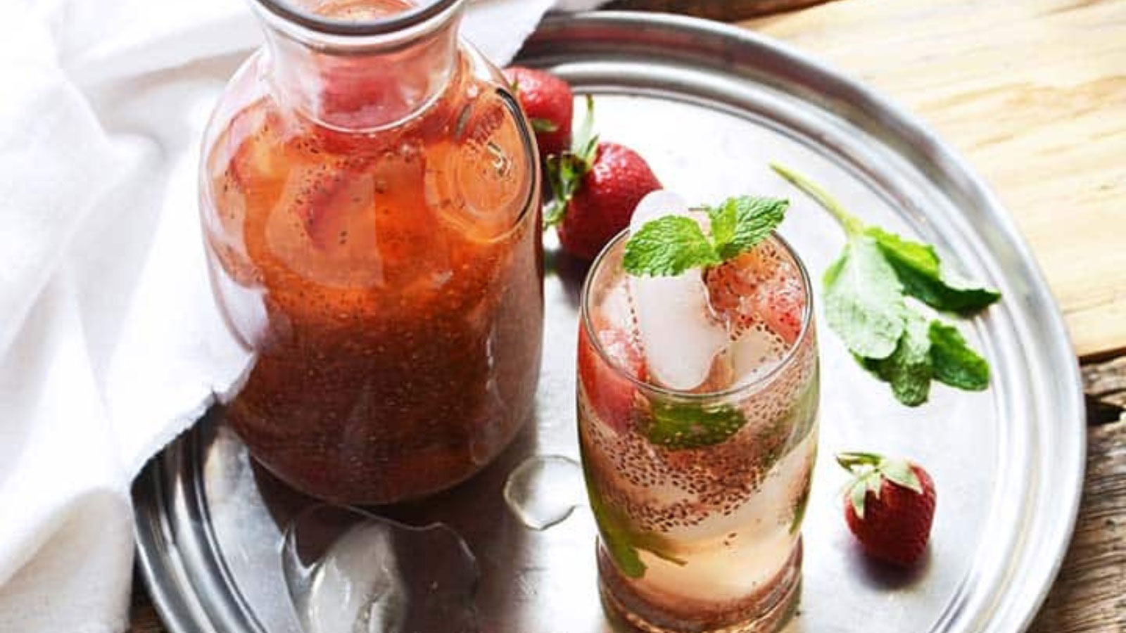 A glass and pitcher filled with chia seed green tea with fresh strawberries, sitting on a silver tray.