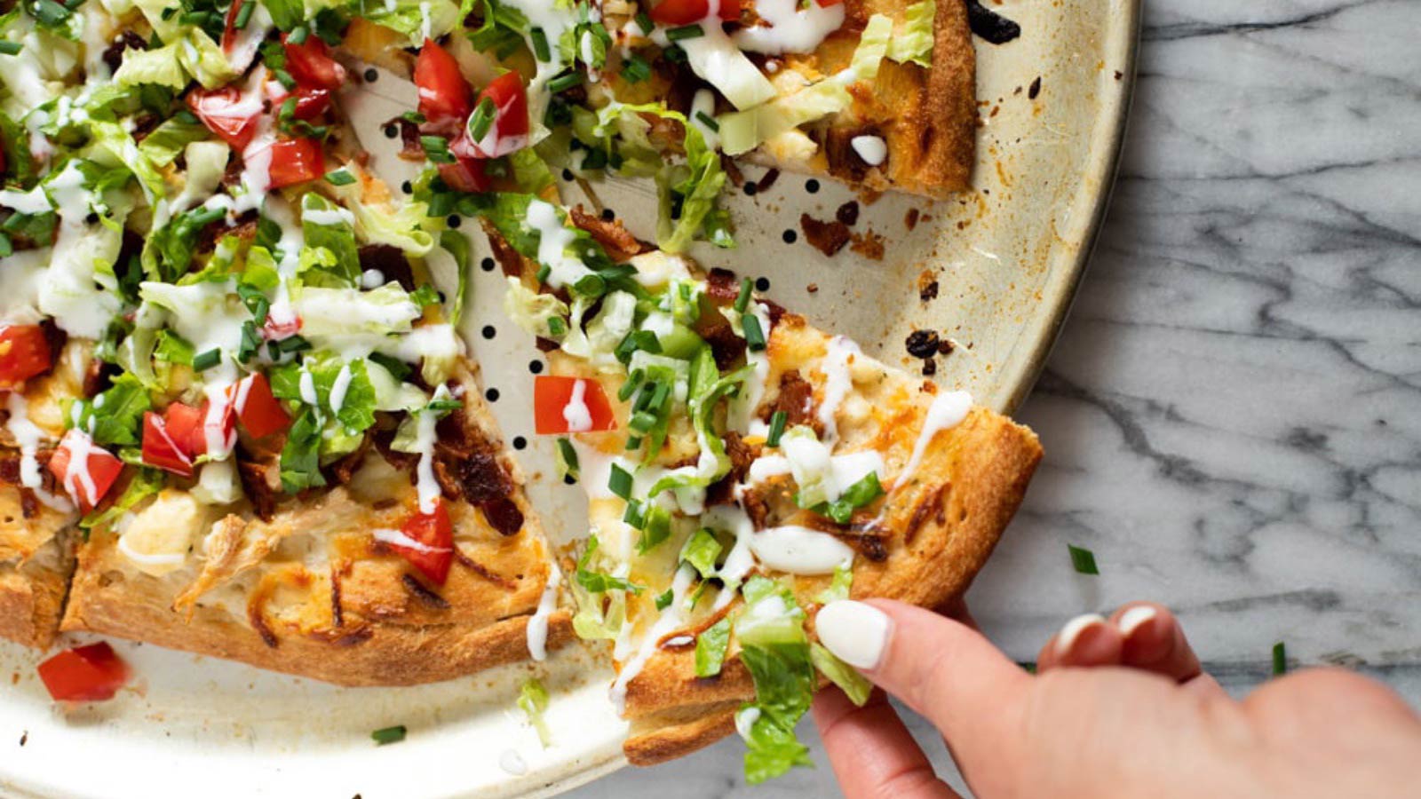 A hand pulls a slice of Chicken Bacon Ranch Pizza from a whole pizza.