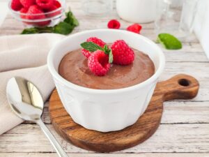 A white bowl filled with raspberry chocolate mousse and garnished with fresh raspberries and mint leaves sits on a table with a spoon next to it.