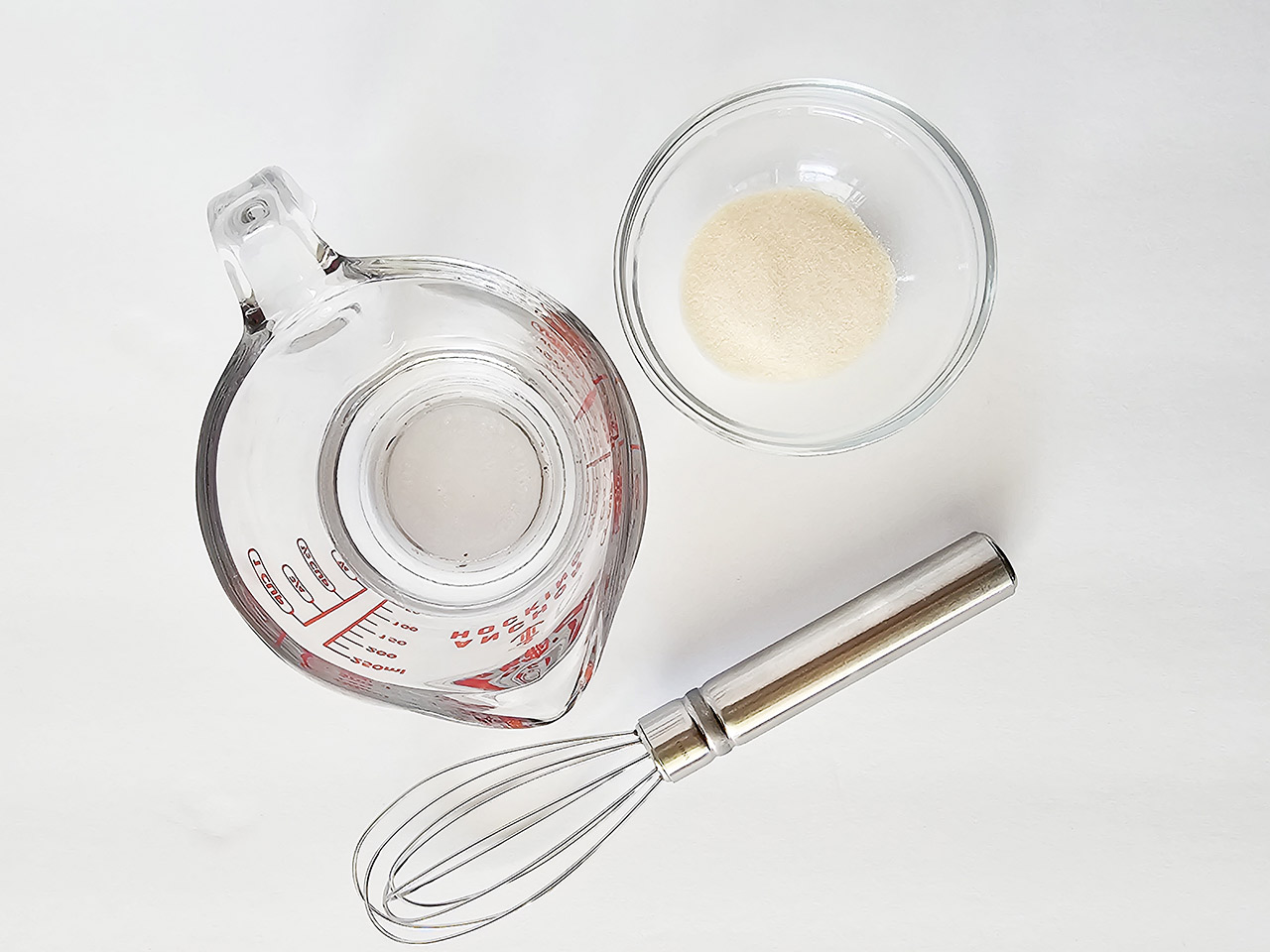 Gelatin powder, water and a whisk on a white surface.