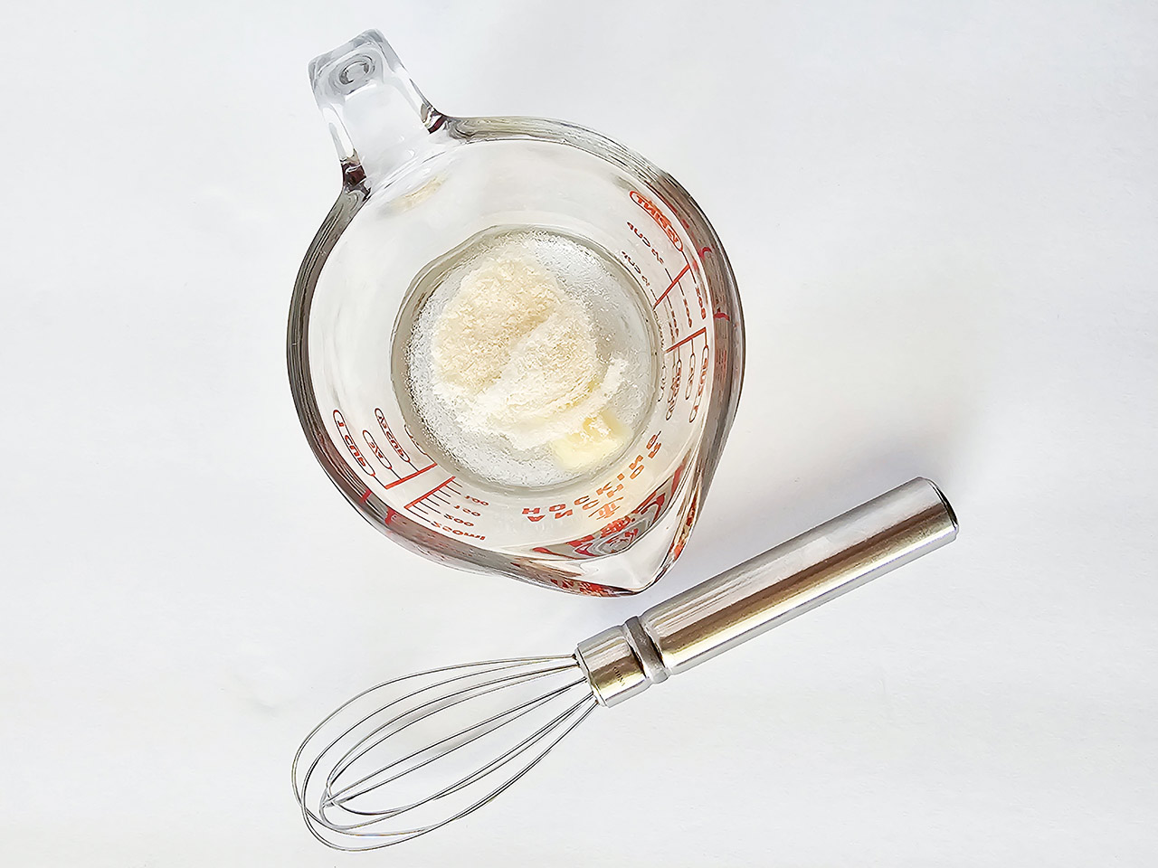 Gelatin powder in hot water in a measuring cup with a whisk laying next to it on a white surface.