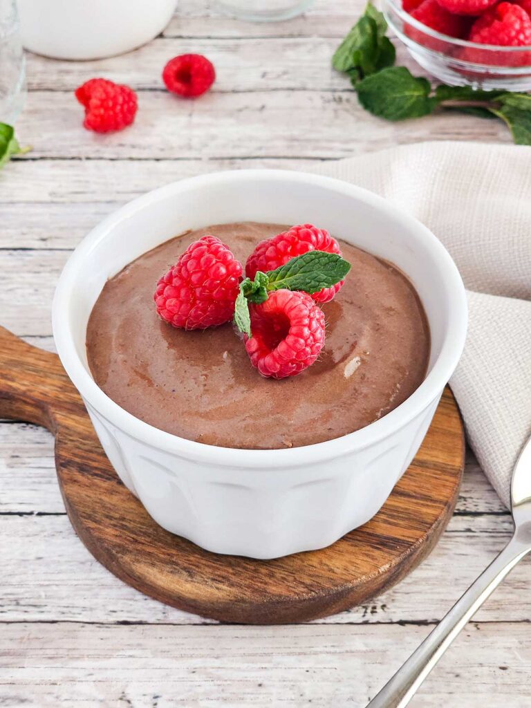 A white bowl on a wood tabletop. The bowl is filled with raspberry chocolate mousse and garnished with three fresh raspberries and a sprig of fresh mint leaves.