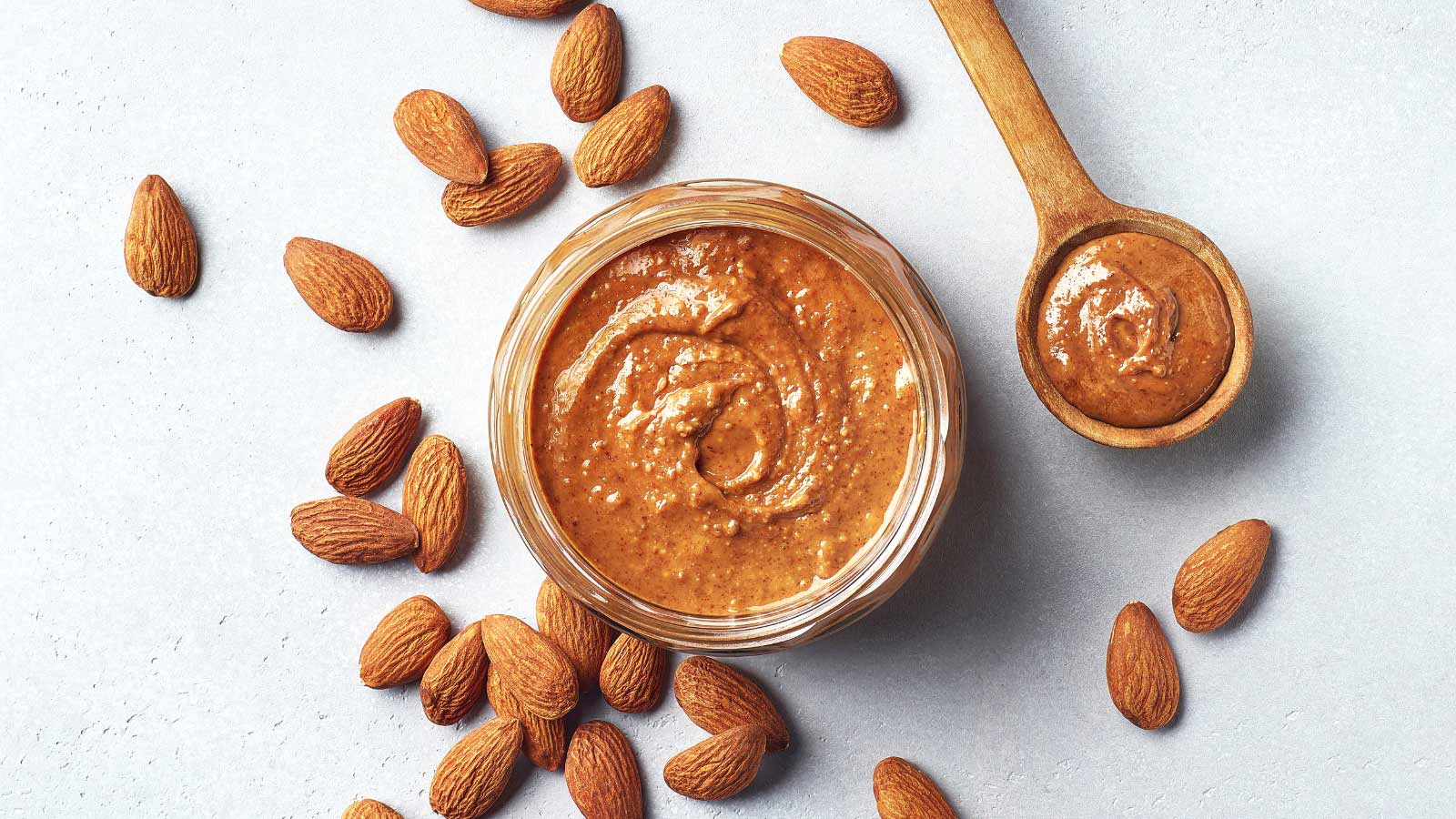 Glass jar and spoon of cinnamon almond butter on light gray background, top view, above
