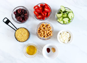 Greek Orzo Salad Recipe ingredients in individual bowls on a white marble surface.