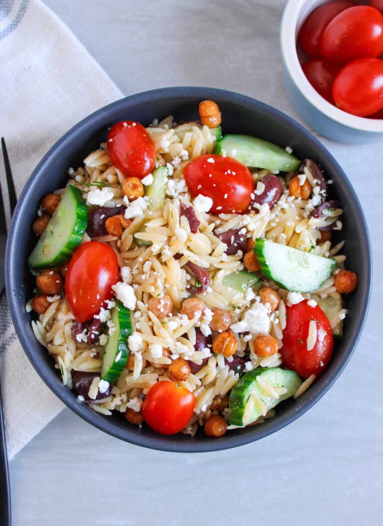 Overhead view of a black bowl filled with Greek Orzo Salad. A black fork lays to the left.