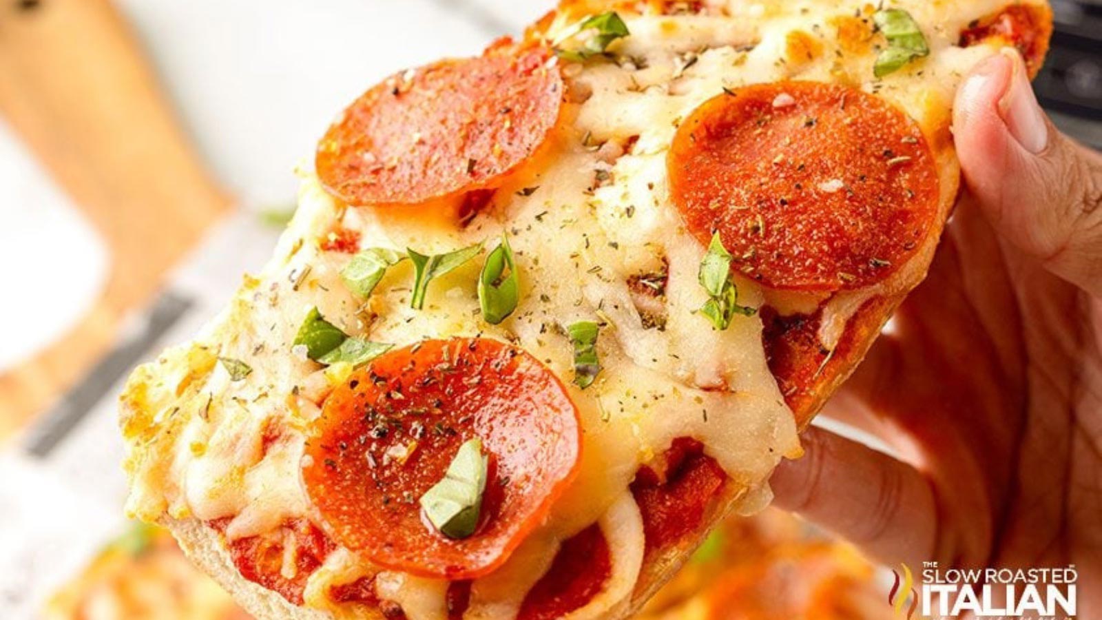 A hand holds a slice of French bread pizza.