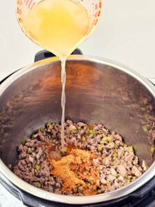 Pouring broth into an Instant Pot with browned meat, spices and vegetables.