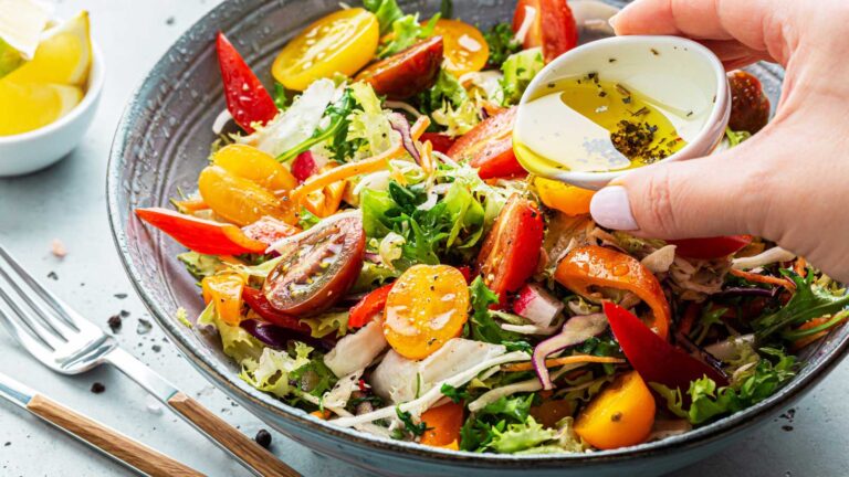 Fresh colorful spring vegetable salad with cherry tomatoes and sweet peppers in the blue bowl. Cook’s hand pouring olive oil with herbs (dressing). Healthy organic vegan lunch or snack close up.