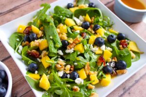 A white platter on a wood surface, holds a Mango Blueberry Salad.