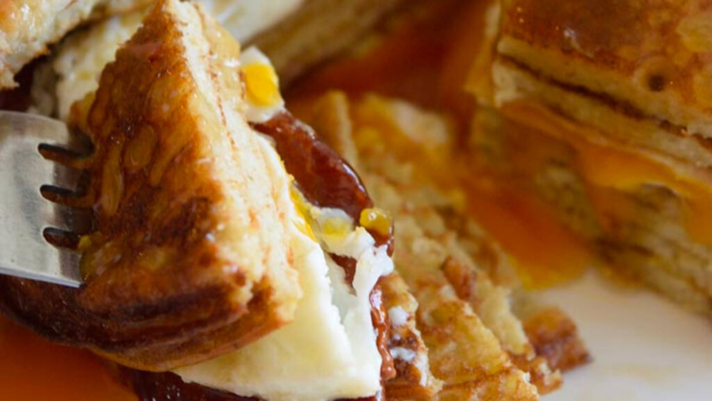 A fork lifts a bite of pancake sandwich off a plate.