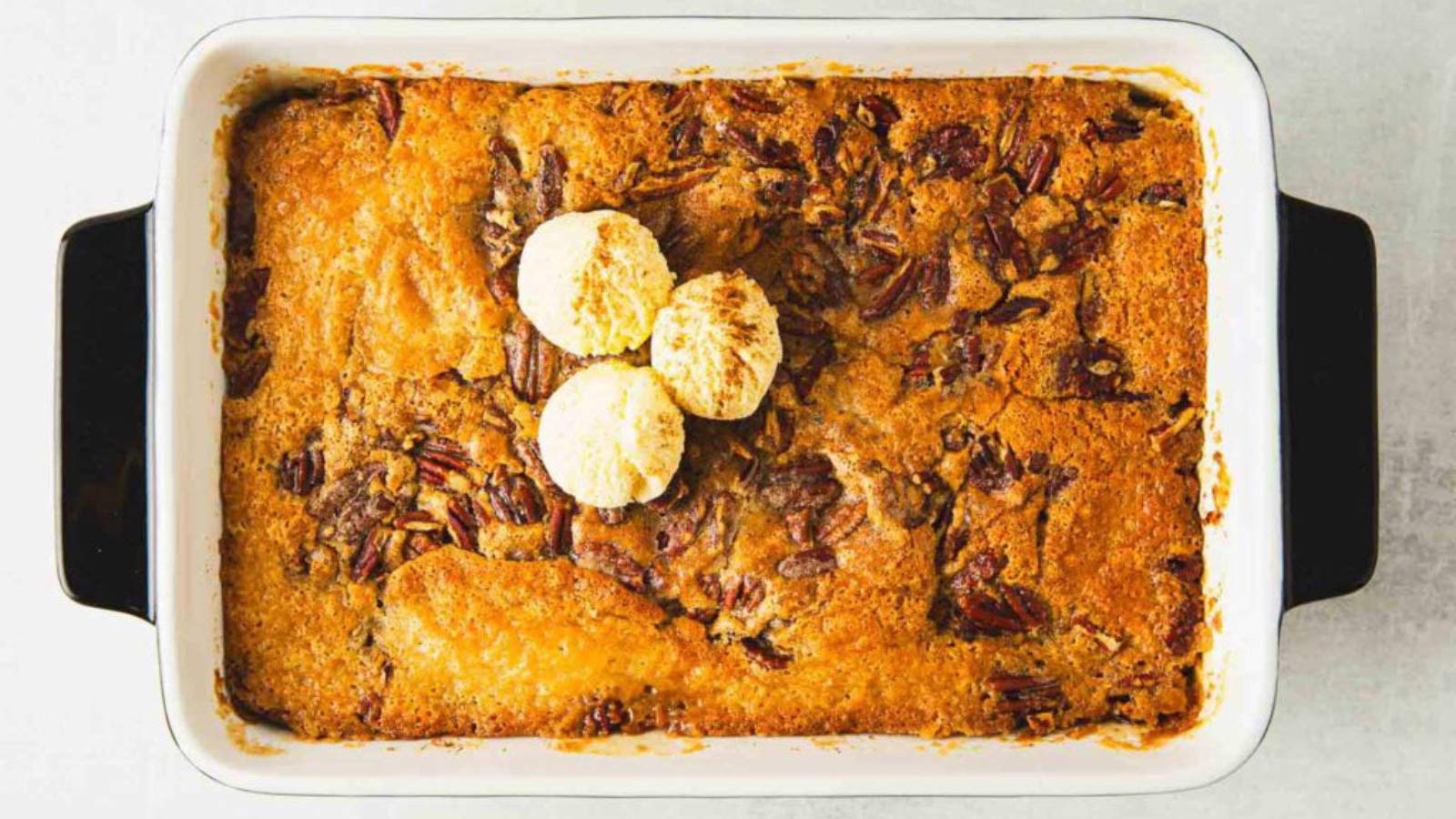 An overhead view of a pecan cobbler in a casserole dish with three scoops of vanilla ice cream in the middle.