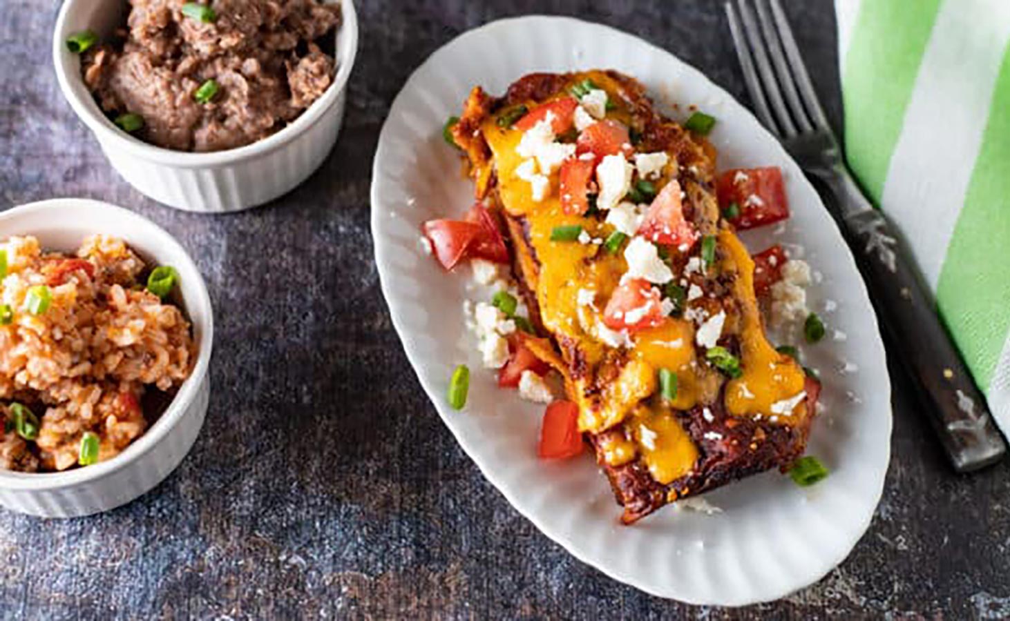 An overhead view of pork enchiladas on a white, oval plate with sides of beans and rice in separate bowls.