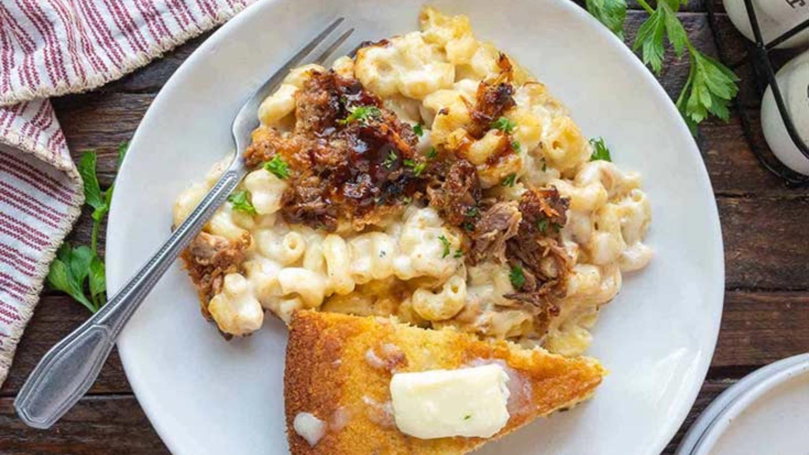 A white plate with a serving of pulled pork mac and cheese. A slice of cornbread lays next to it with a pat of melting butter on top.