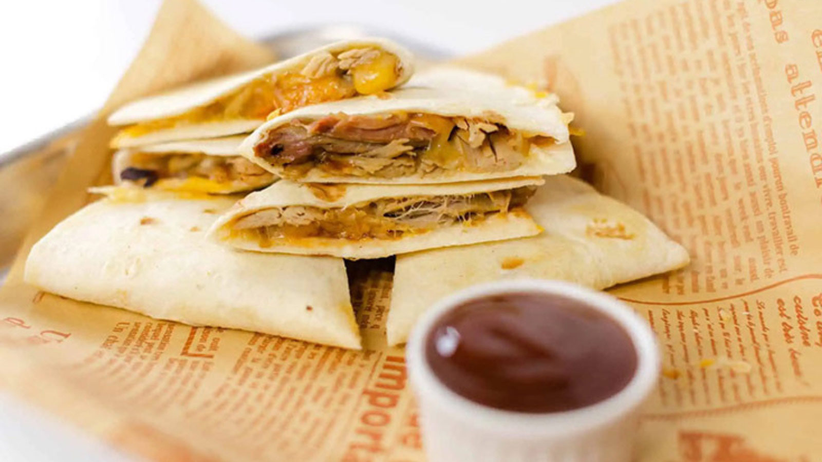 A cut, pulled pork quesadilla lays on parchment next to a small white bowl of dipping sauce.